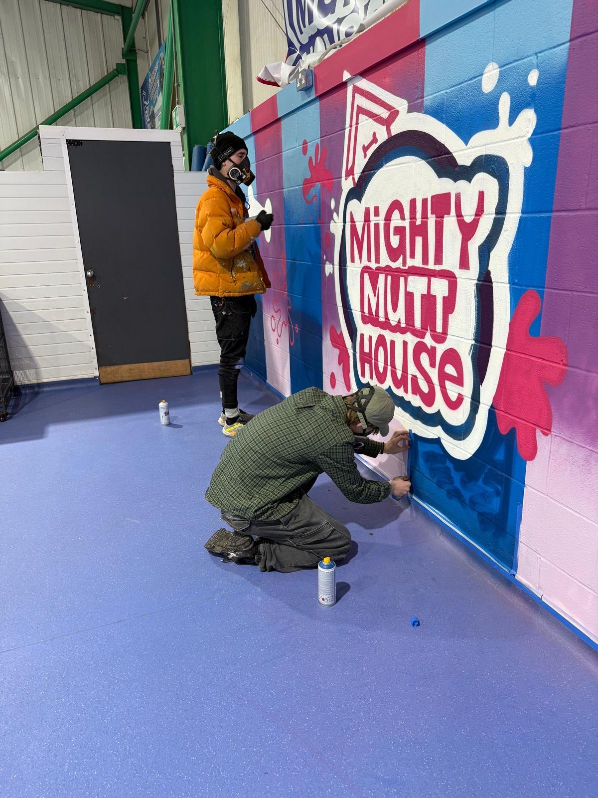 Two spray painters at work on the Mighty Mutt House logo on the arena wall, wearing masks, one standing and one kneeling with spray cans on the floor