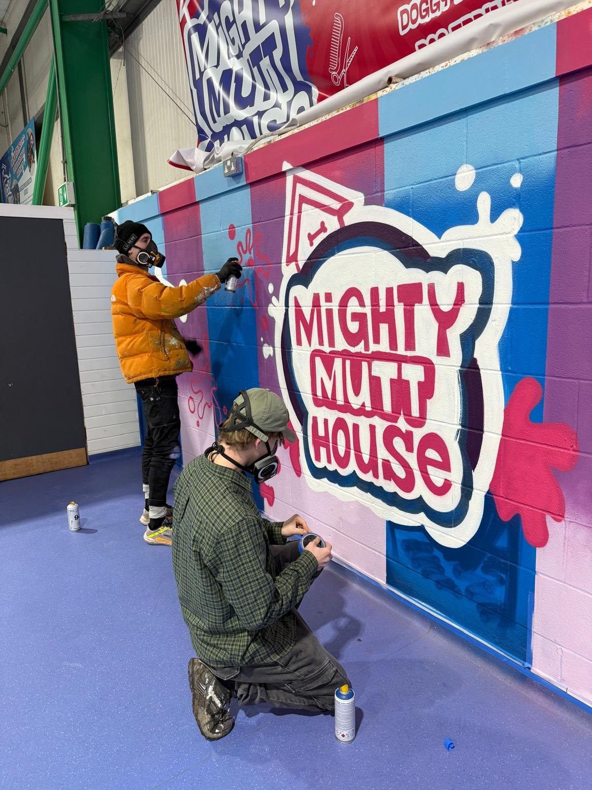 A spray painter in a mask finishing the outline of the painted Mighty Mutt House logo while a second painter kneels beside the wall